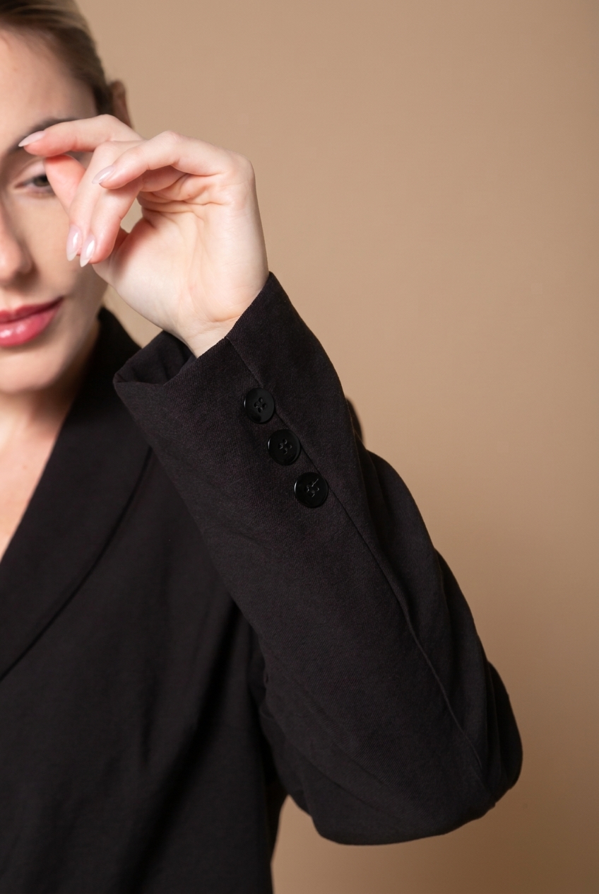 Detalhe da manga e dos botões de um blazer preto Bozzetti, com modelo sobre fundo marrom